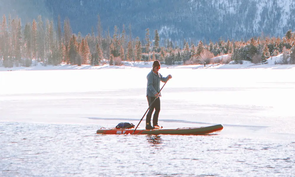 Bild von Person auf SUP Board in Rot im Winter angezogen mit warmer Winterjacke, Brille und Haube - Hintergrund Schnee Berge