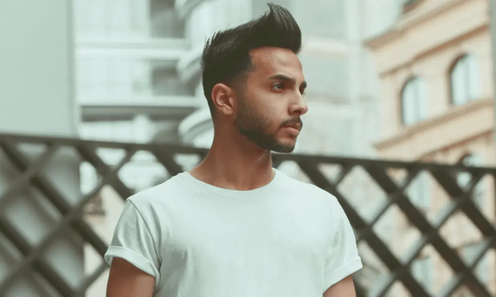 Bild von Mann mit schwarzen Haaren und Foehnfrisur schaut nach rechts und weissem T-Shirt mit Häuserhintergrund